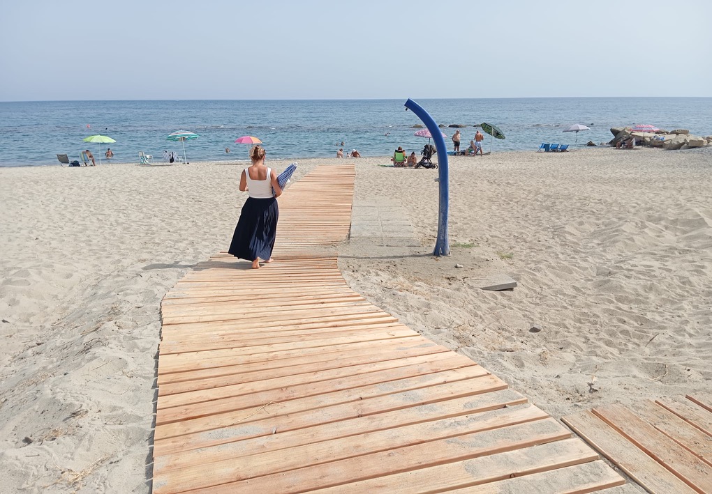 Passerella in legno sulla spiaggia che conduce verso la riva, percorsa da una donna con un ombrellone chiuso; intorno si trovano bagnanti, ombrelloni, sedie da spiaggia e una doccia all’aperto.