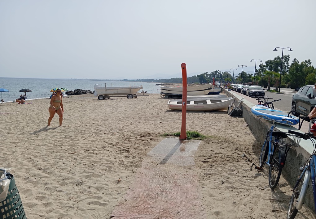 Accesso alla spiaggia con doccia all’aperto, piccole imbarcazioni sulla sabbia e biciclette appoggiate al muretto; una bagnante cammina verso la riva, mentre sullo sfondo si vedono persone in mare, il lungomare e le montagne.