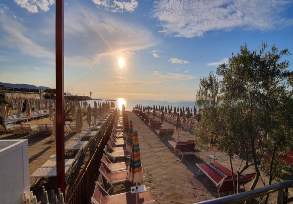 Stabilimento balneare al tramonto, con file di lettini e ombrelloni disposte lungo la spiaggia e il sole che si riflette sul mare.