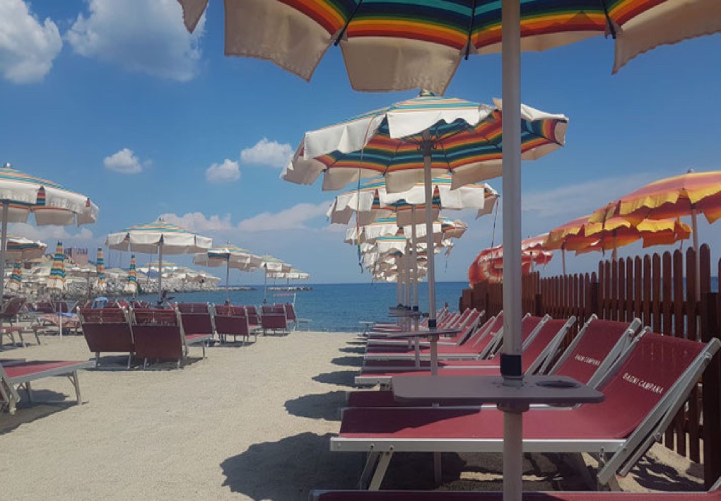 Spiaggia attrezzata con numerosi lettini e ombrelloni, un ampio passaggio centrale e il mare sullo sfondo.
