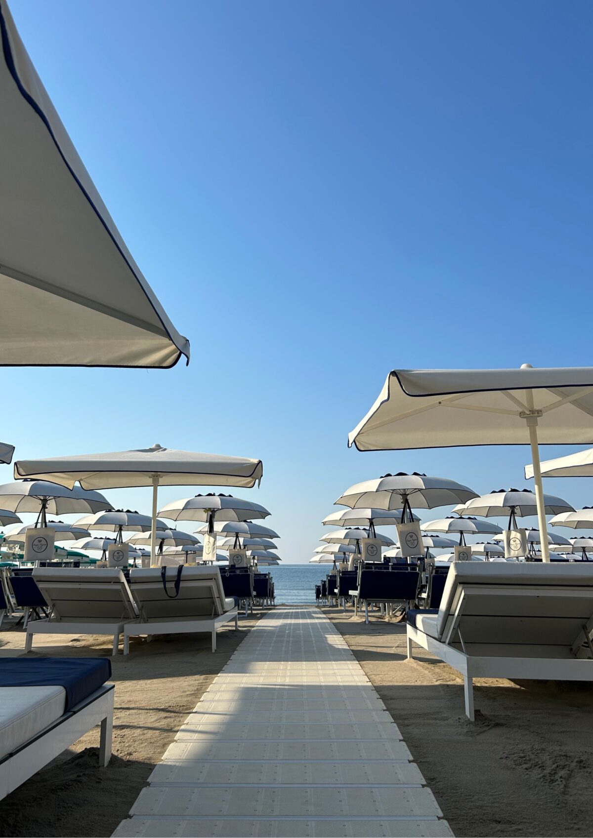 Vista simmetrica in prospettiva di una spiaggia attrezzata: al centro si sviluppa una passerella rigida che attraversa file ordinate di postazioni con ombrelloni e lettini, creando un corridoio d'accesso che conduce direttamente verso la linea d'orizzonte del mare.