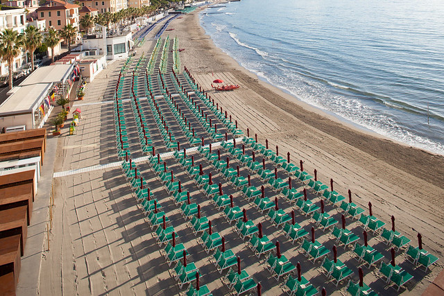 L'immagine mostra una vasta estensione di spiaggia organizzata con file fitte e lunghe di ombrelloni chiusi e lettini allineati simmetricamente sulla sabbia, che si estendono parallelamente alla linea del mare. Sul lato sinistro, una struttura coperta a più moduli si affaccia sulla spiaggia, mentre un percorso pavimentato corre lungo il perimetro dello stabilimento balneare. Il mare e la linea costiera sullo sfondo definiscono l'area.