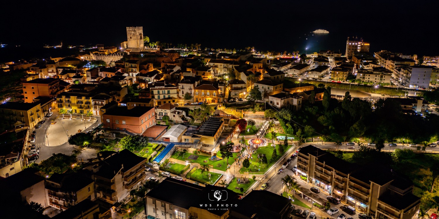 - Veduta aerea notturna di Brolo, con il centro abitato illuminato, la Villa Comunale, il castello medievale e il mare sullo sfondo.