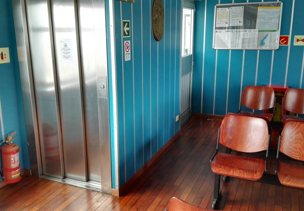 dettaglio dell'interno: la porta dell'ascensore si trova a fianco di alcune file di sedie color legno in una sala chiusa, con pareti pannellate e pavimento in legno.