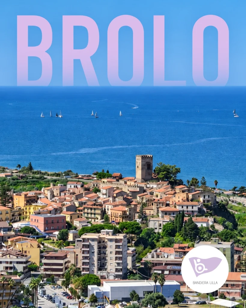 Vista panoramica di Brolo, in Sicilia, con il centro abitato affacciato sul mare, il castello medievale in posizione sopraelevata e alcune barche a vela al largo. In alto compare la scritta “BROLO” e in basso a destra il logo di Bandiera Lilla.