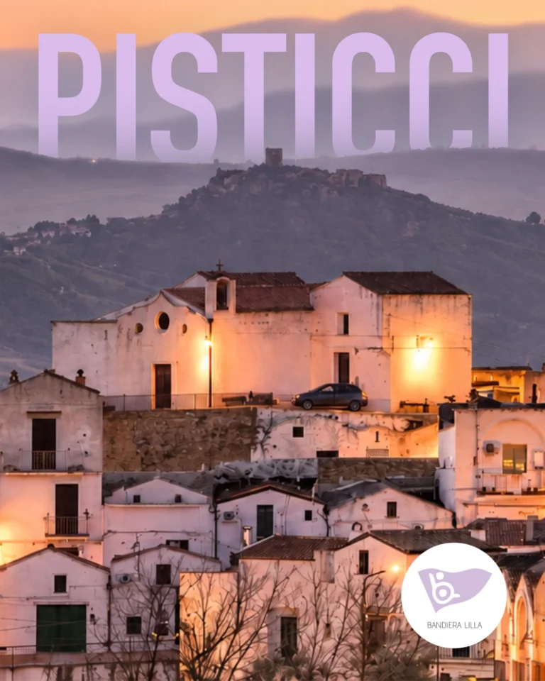 Veduta di Pisticci al tramonto, con le case bianche del borgo illuminate e le colline sullo sfondo. In alto compare la scritta “PISTICCI” e in basso a destra il logo di Bandiera Lilla.