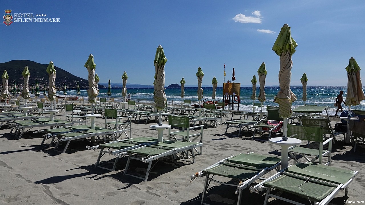 Panoramica di una spiaggia attrezzata con file di lettini, sedie e ombrelloni chiusi disposti direttamente sulla sabbia. Sulla destra è presente una postazione di salvataggio rialzata, mentre sullo sfondo si vedono il mare e un promontorio costiero.