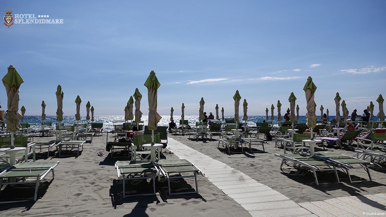 Spiaggia attrezzata divisa da una passerella rigida centrale a raso sabbia. Ai due lati del percorso si trovano file ordinate di lettini e ombrelloni chiusi che conducono verso la riva del mare.