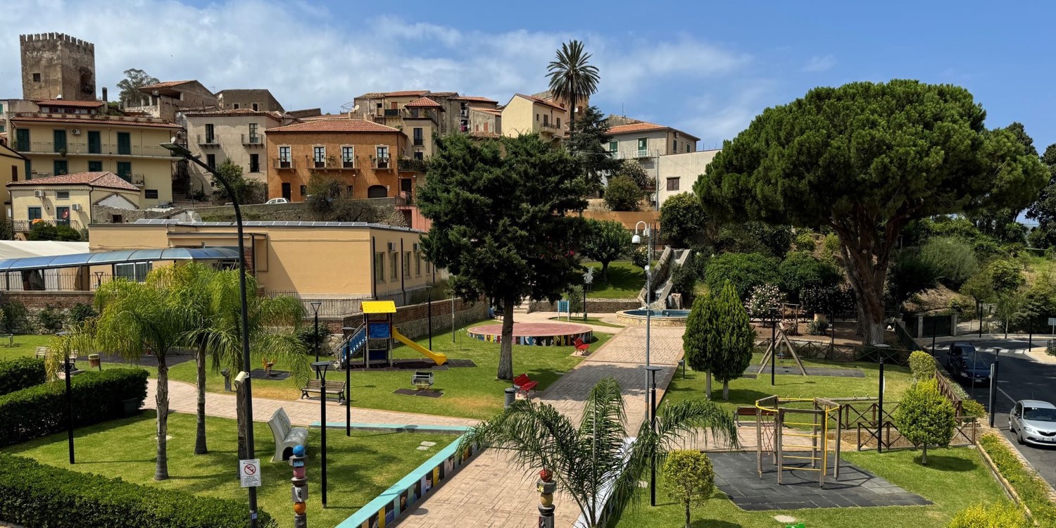Vista panoramica della Villa Comunale di Brolo, con prati, alberi, vialetti e aree gioco; sullo sfondo si distinguono il borgo e la torre del castello.